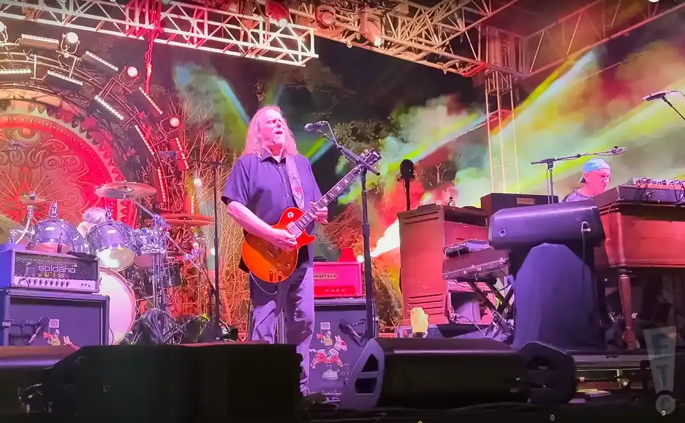 A guitarist plays on a brightly lit stage with drums and keyboards.