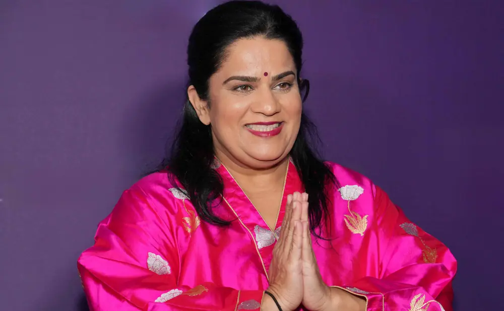 A woman in a pink robe with embroidered flowers smiles and places her hands together in a greeting.