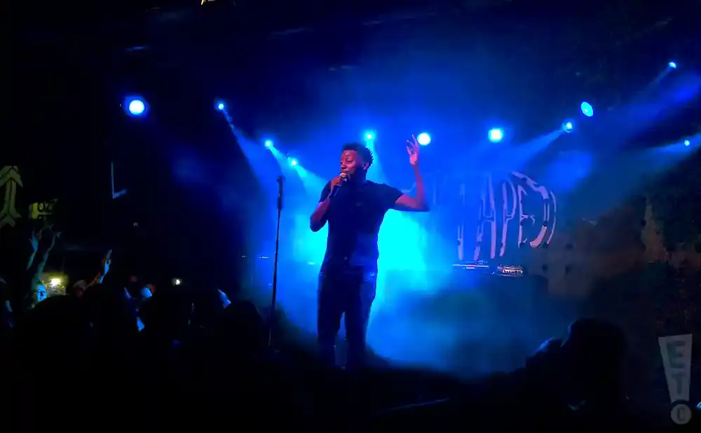 A male performer sings into a microphone on a stage bathed in blue light, with a crowd visible in the foreground.