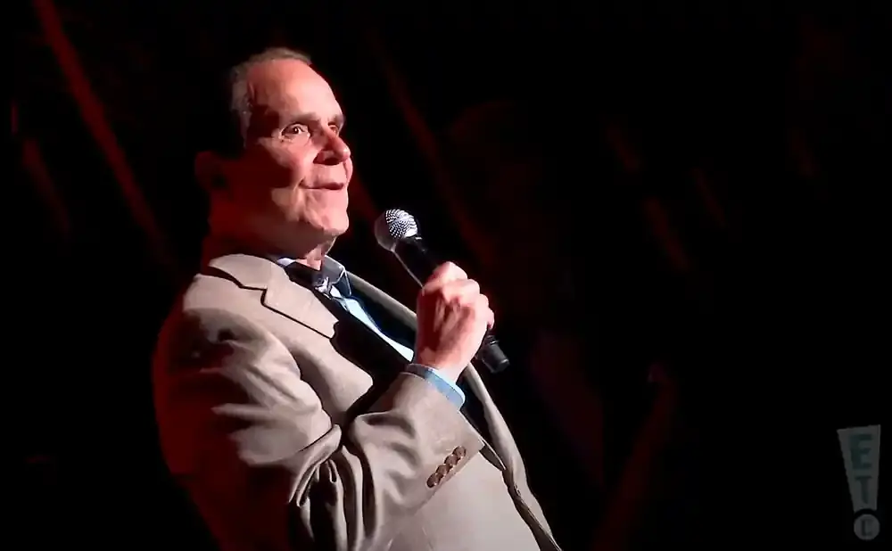 A man in a light-colored suit and blue shirt holds a microphone, looking to his left.