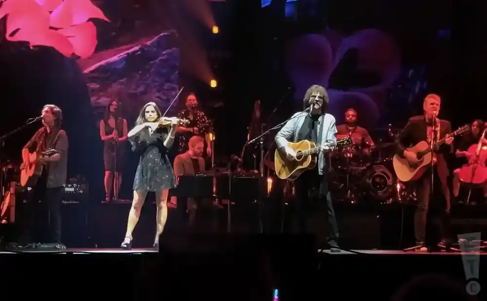 A band performs on stage, with a violinist in the foreground and guitarists and a drummer behind her.