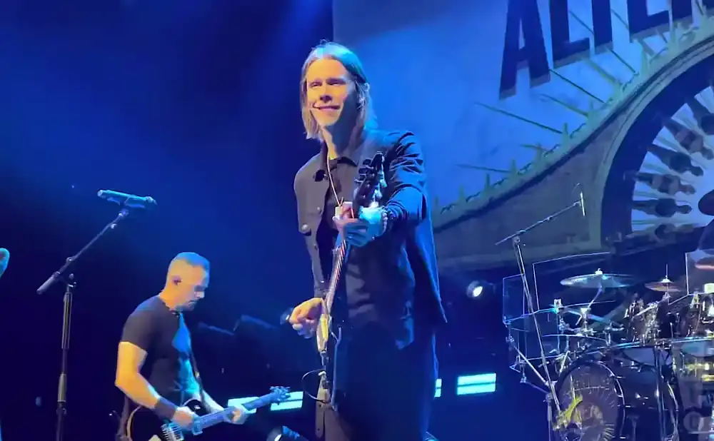 A guitarist smiles and plays a black electric guitar on a stage with blue lighting.