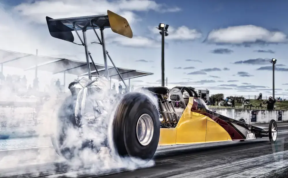 Dragster doing a burnout, kicking up smoke, on a track with spectators in the background.