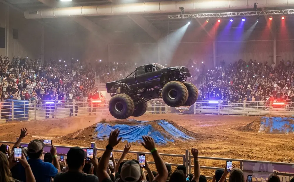 Monster truck jumps over a dirt mound as a large crowd watches with their phones out.