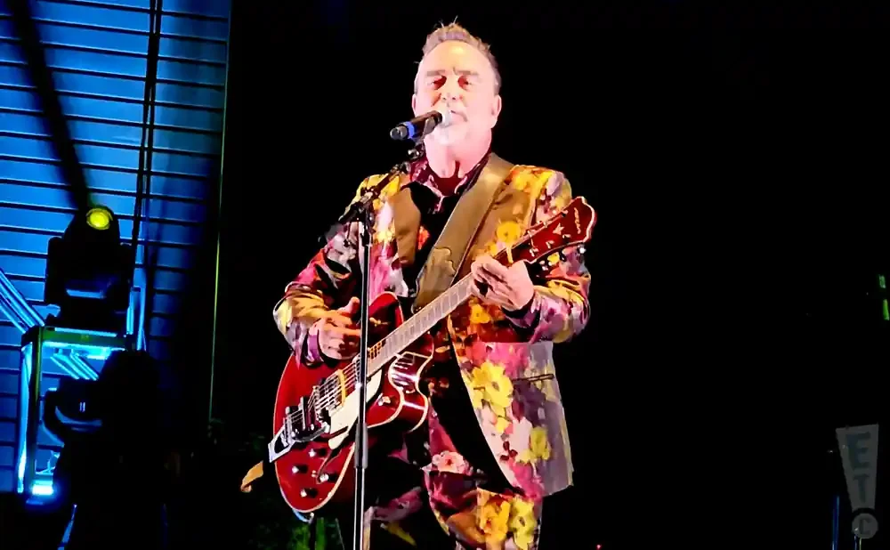 Man in a colorful floral suit plays a red guitar and sings into a microphone on stage.