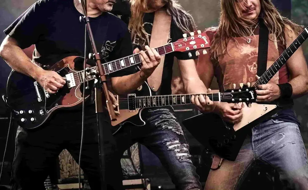 Three men play guitars on stage, two with long hair and ripped jeans, the third in a black shirt.