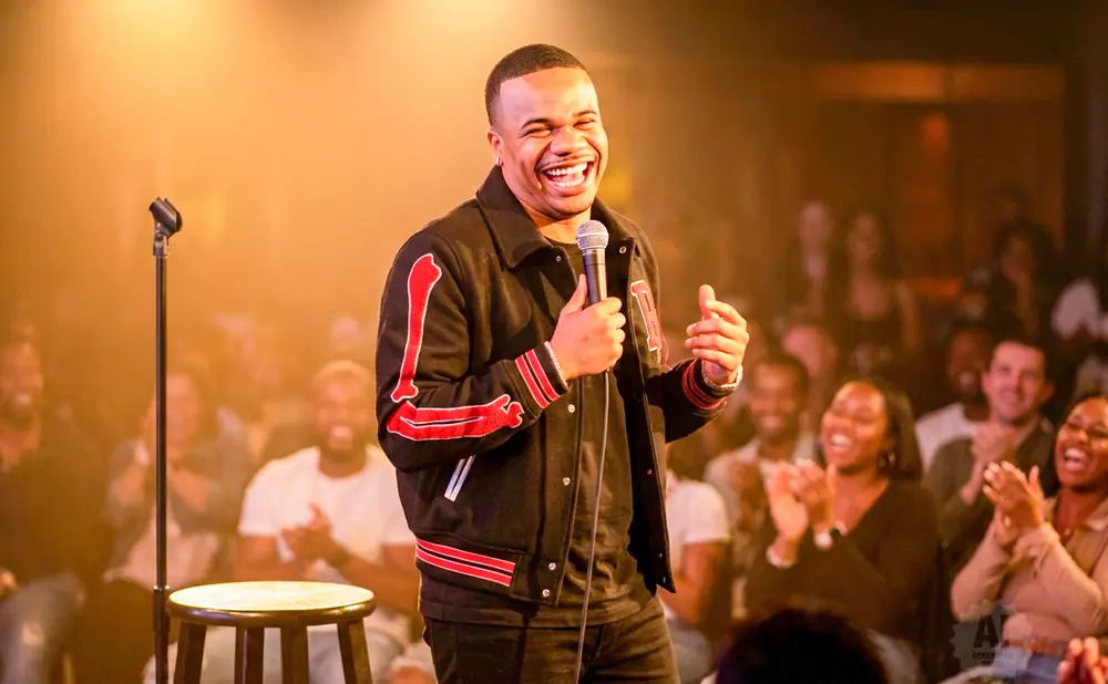 A comedian laughs on stage, holding a microphone, with a laughing audience in the background.