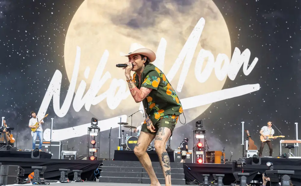 A male singer in a cowboy hat and orange fruit-print shirt performs on stage, holding a microphone.