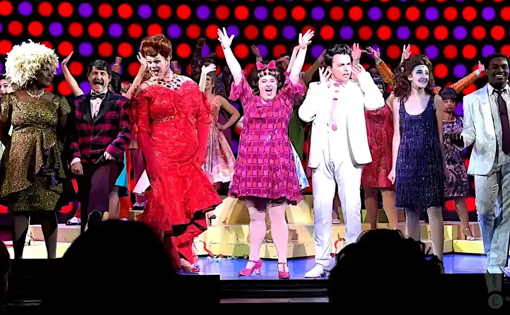 A cast in colorful costumes on stage, with a bright red and purple dotted backdrop.