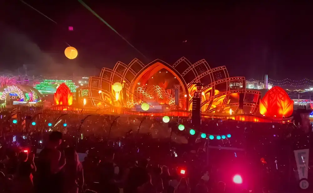 A massive outdoor stage at night with a large crowd in front. The stage is lit with orange and red lights and decorated with lotus flower-like structures.