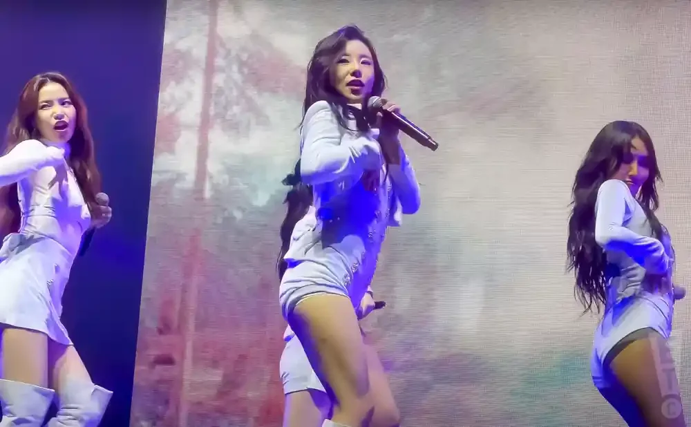 Three women in white outfits perform on stage, with the center singer holding a microphone.