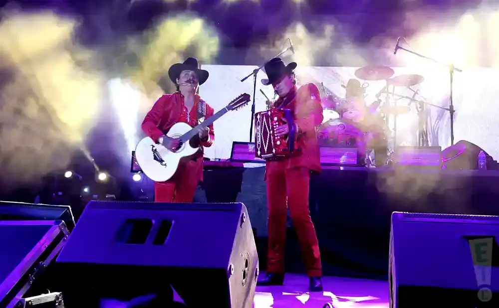 Musicians in red suits and cowboy hats perform on stage with a guitar and accordion, lit by stage lights and smoke.