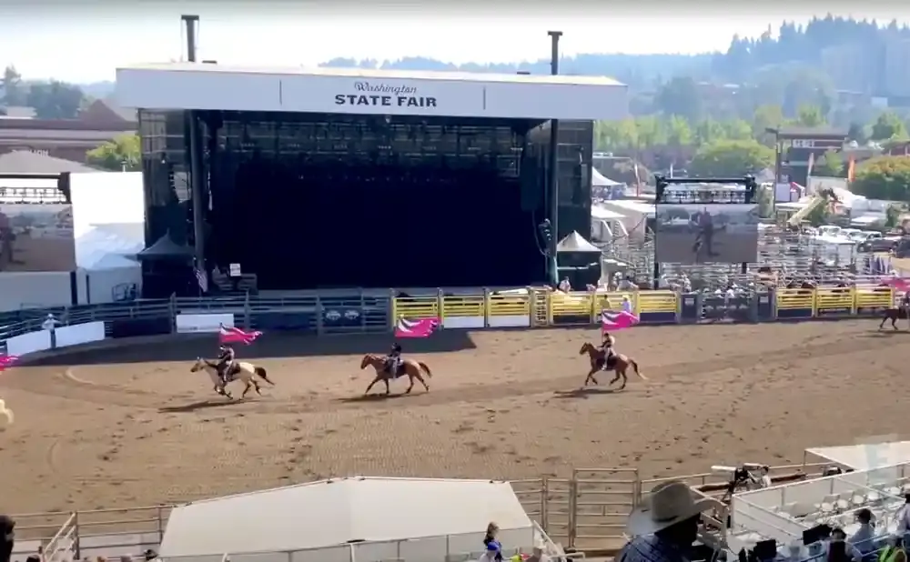 Horse riders with flags race in a rodeo arena in front of a large stage at the Washington State Fair.