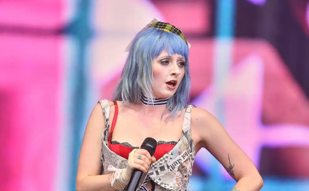 Singer with blue hair and newspaper outfit performing on stage with a pink and blue background.