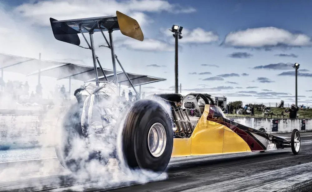 A yellow and red dragster with a large rear wing kicks up smoke as it races down a track.
