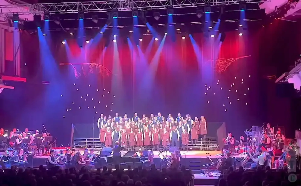 A choir and orchestra perform on a stage with red and blue lighting and decorative branches.