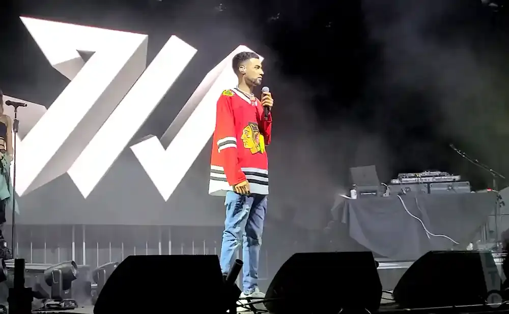 A performer in a red hockey jersey stands on a stage with a large white logo behind him.