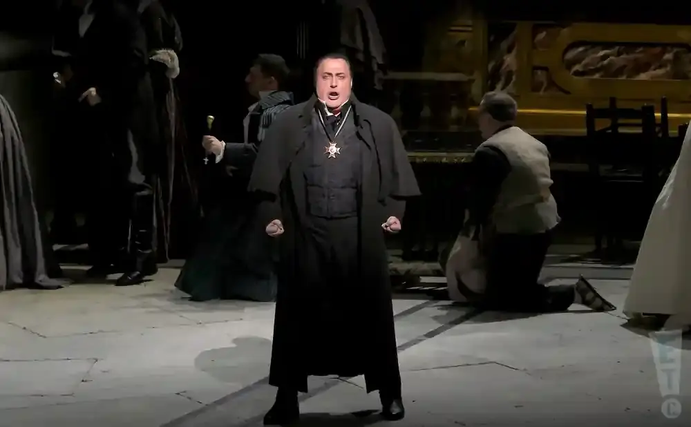 Opera singer in a black robe and medal sings with fists clenched.