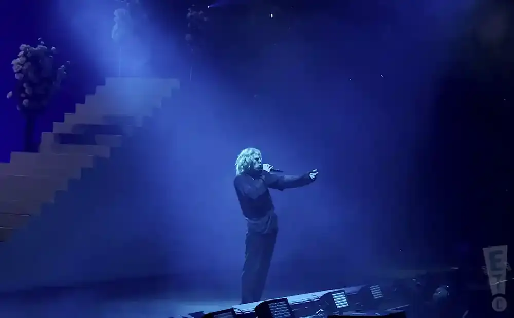 A singer performs on a stage with blue lighting and smoke, with a staircase behind them.