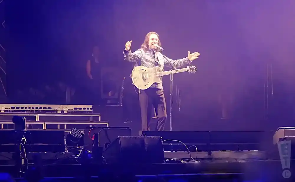 A male guitarist sings and plays an electric guitar on a brightly lit stage.