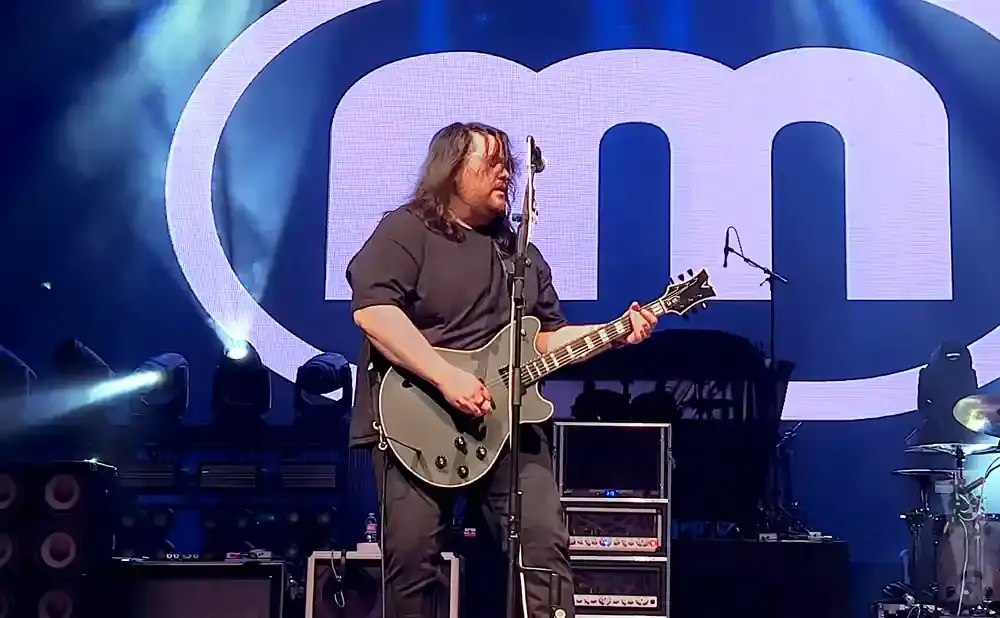 A man plays a gray electric guitar on stage with large 