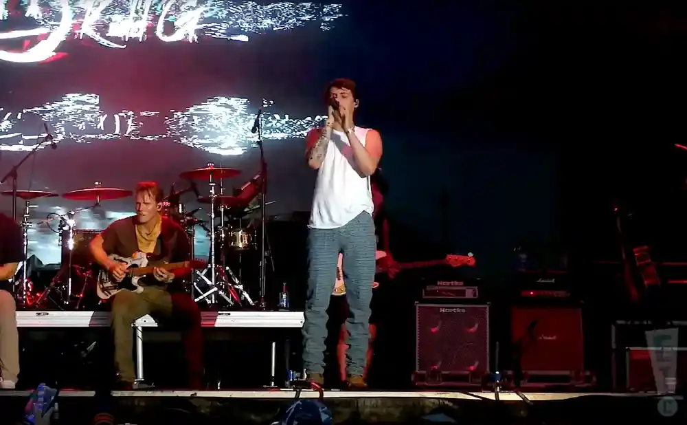 A male singer in a white shirt and patterned pants performs on stage with a guitarist and drums in the background.