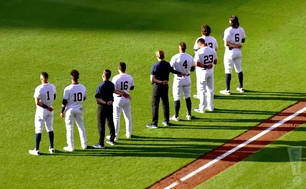 Baseball players in uniform stand in a line on a grassy field, with some wearing numbered jerseys.