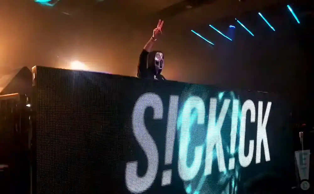 A masked DJ stands behind a large LED screen displaying 
