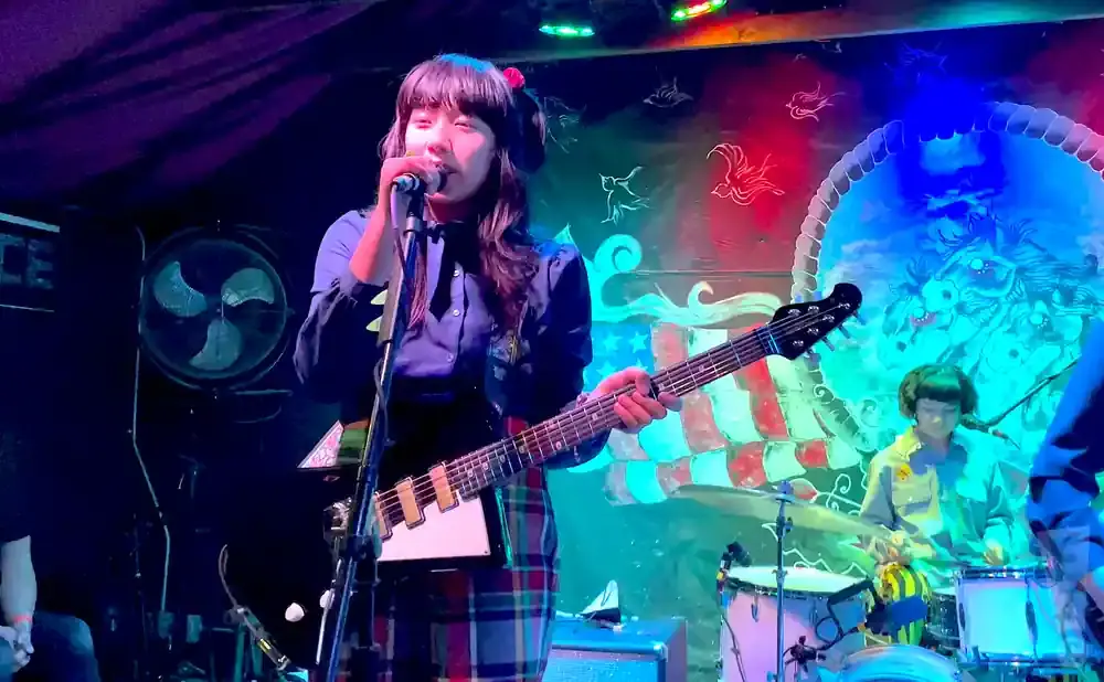 A female bassist sings into a microphone with a guitar. A drummer plays drums in the background.