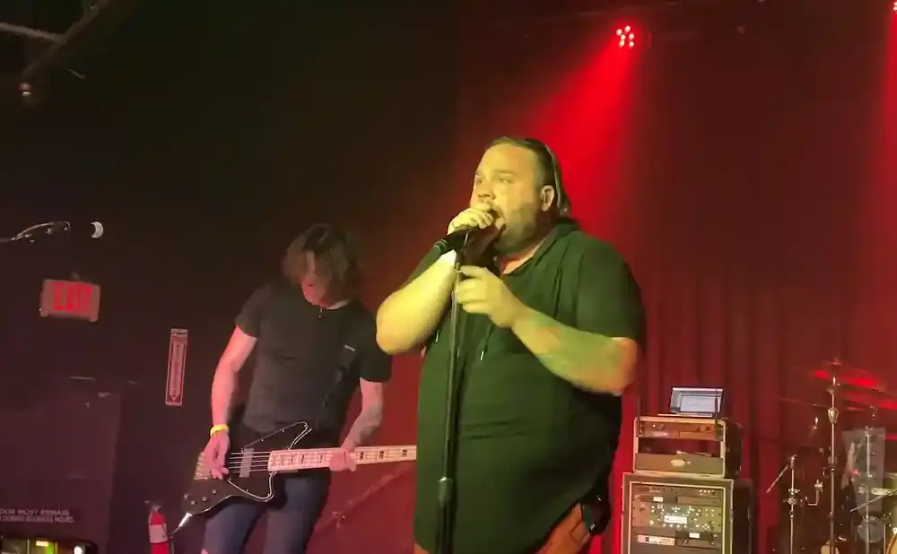 A singer in a black shirt sings into a microphone, while a guitarist plays a bass guitar to his left on a stage with red lighting.