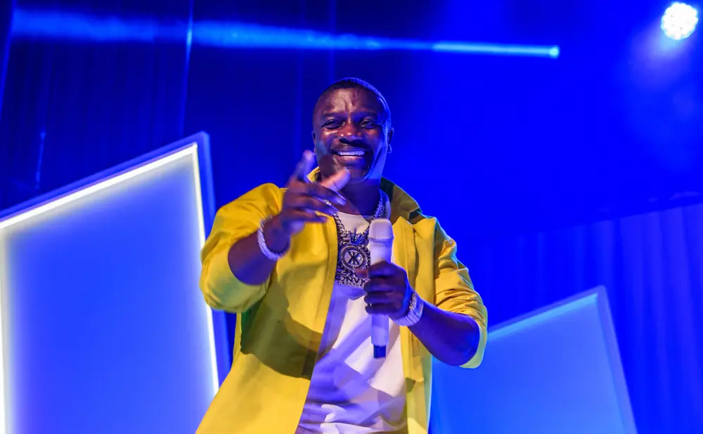 Akon in a yellow jacket smiles and points while holding a microphone.