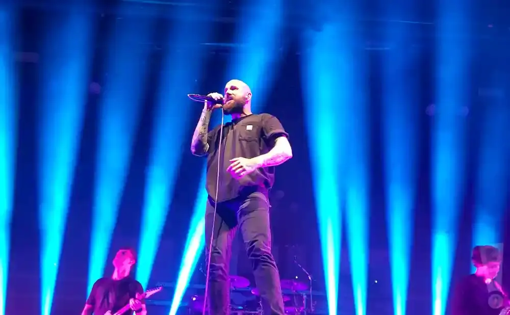 A bald singer in a black t-shirt and jeans sings into a microphone on a stage with blue spotlights.