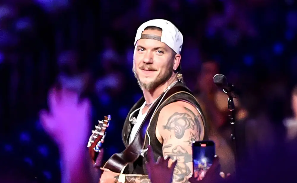 Man with tattoos and a white baseball cap plays guitar on stage with purple lights.