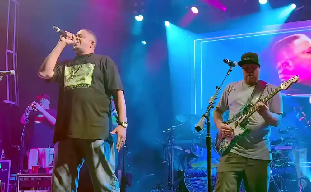 A male rapper performs into a microphone on stage, while a guitarist plays an electric guitar to his right.