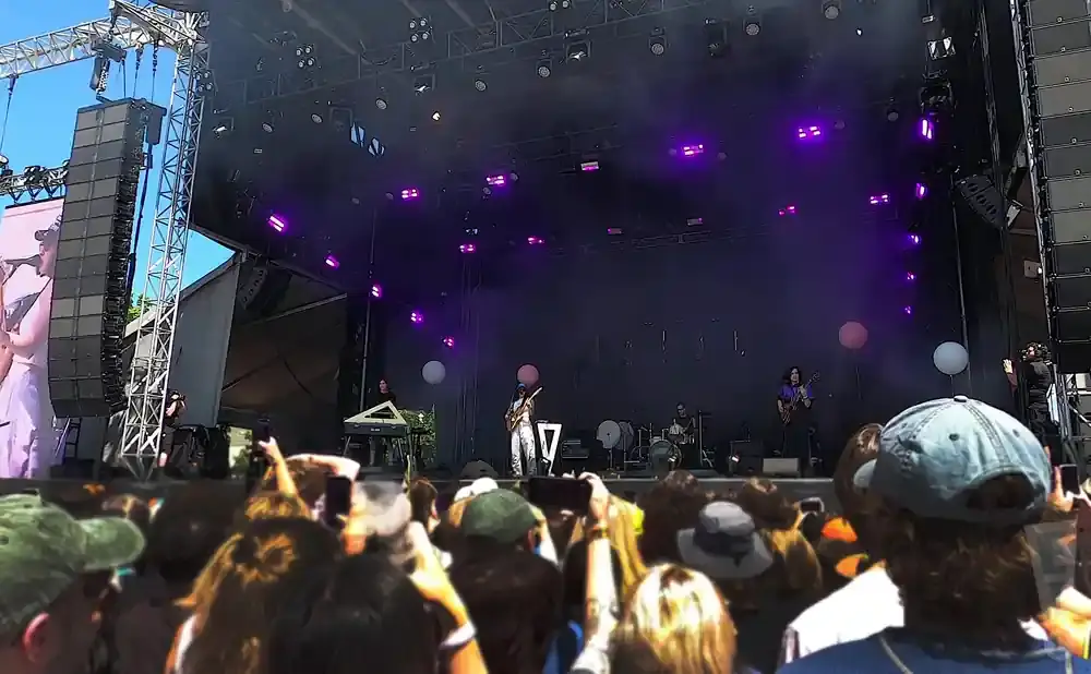 A band performs on a stage at an outdoor concert, with a large crowd in the foreground.