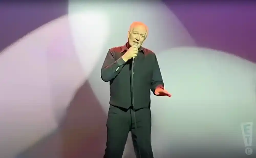 Comedian Bill Burr stands center stage in a black shirt and pants, holding a microphone and gesturing with his right hand.