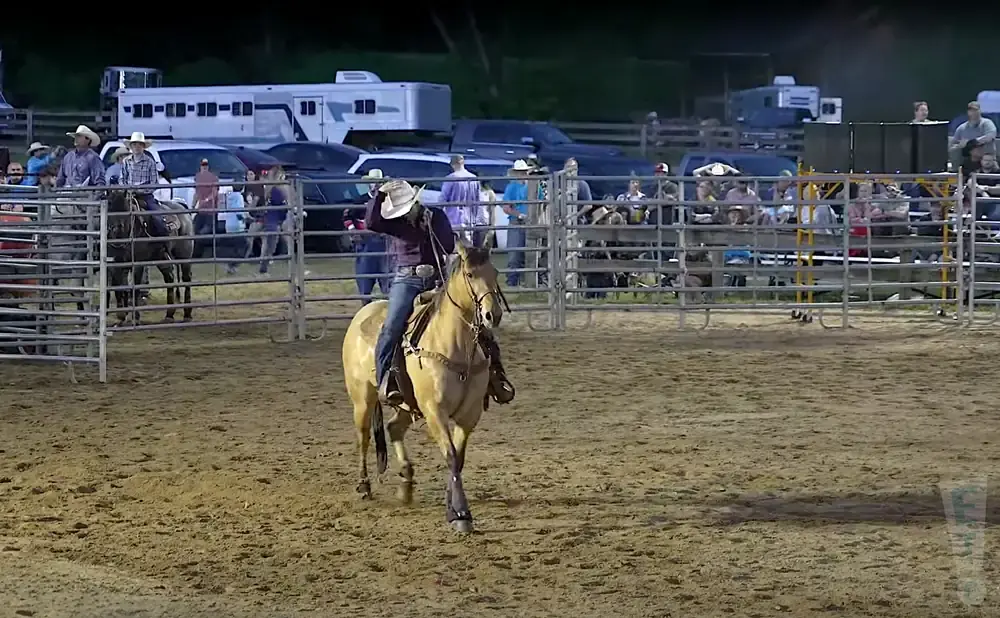 Cowboy on a tan horse in a rodeo arena, with spectators and trailers in the background.