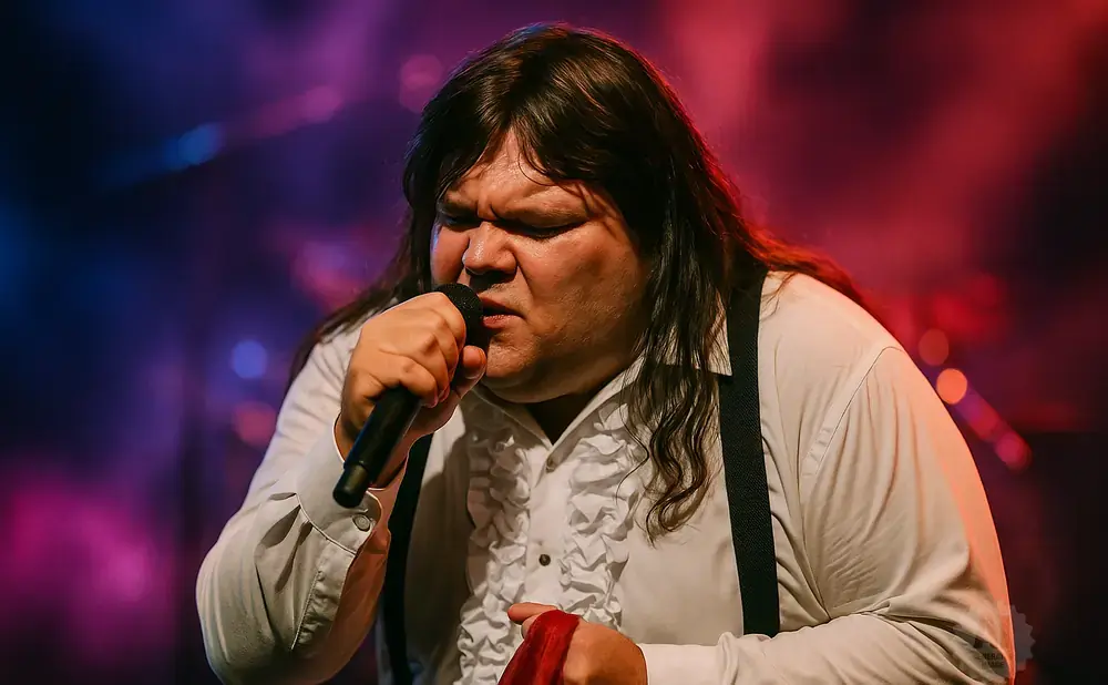 A man with long dark hair sings into a microphone, wearing a white ruffled shirt and black suspenders.