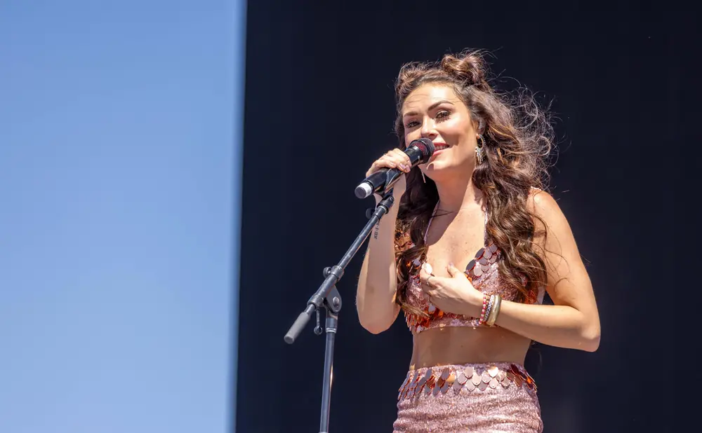 A woman in a sparkly pink outfit sings into a microphone on a stage with a dark background.