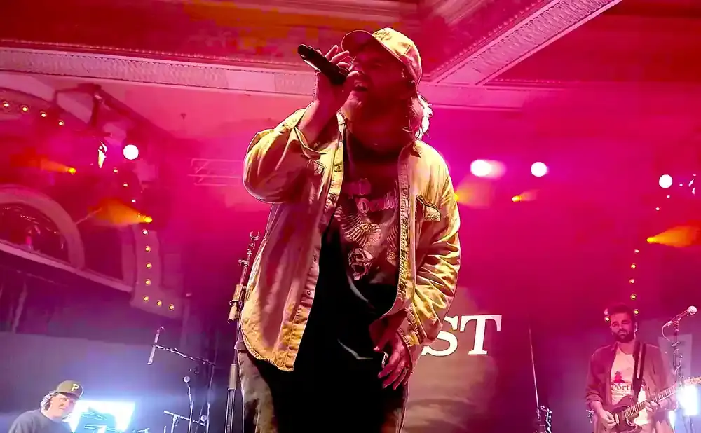 A man in a tan jacket and cap sings into a microphone on a pink-lit stage. Another man plays guitar to his right.