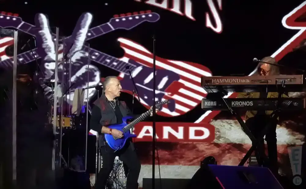 Musicians perform on stage with a backdrop of guitars and an American flag motif.