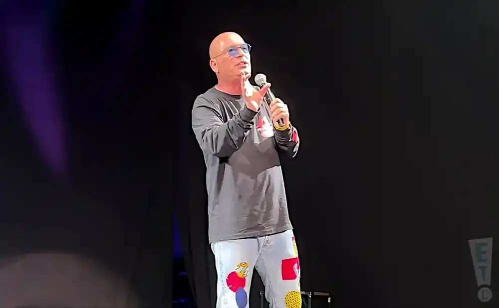 Bald man in a long-sleeved shirt and jeans with colorful patches speaks into a microphone on stage.