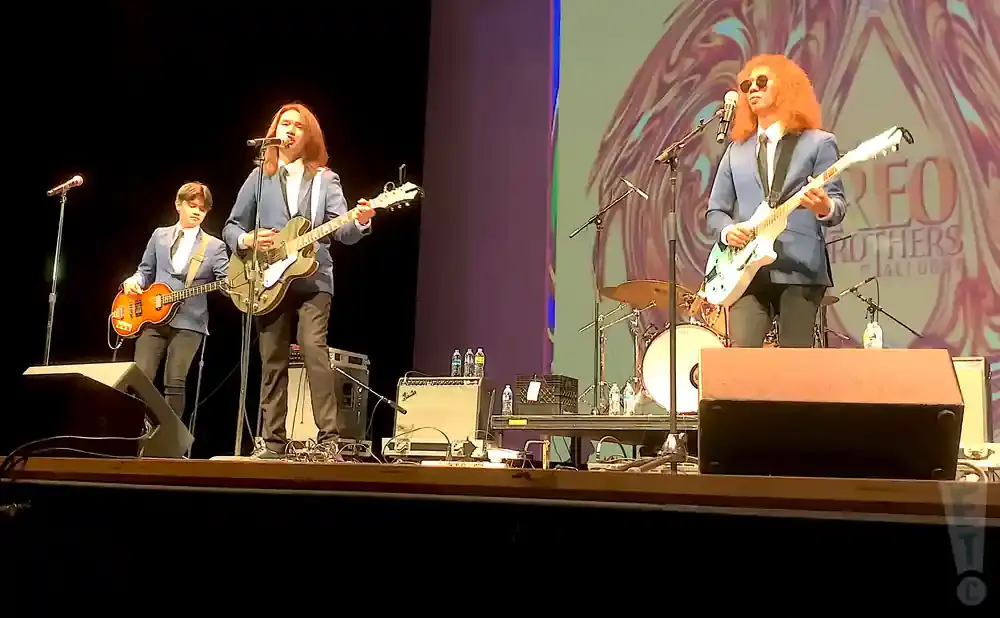 Three men in blue blazers play guitars on stage with drums and amps behind them.