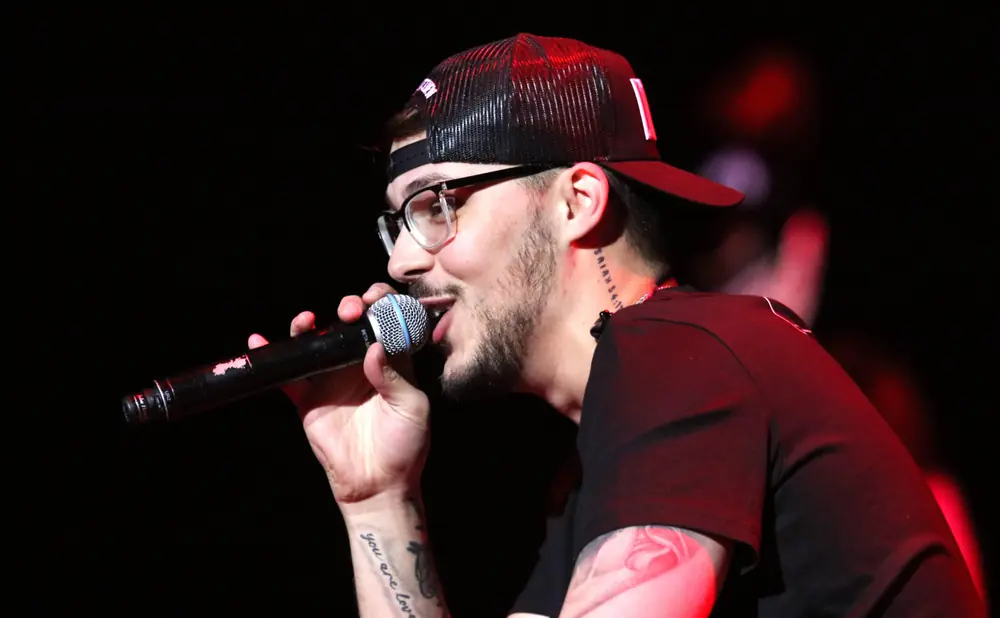 A man with glasses and a backward baseball cap sings into a microphone on stage.
