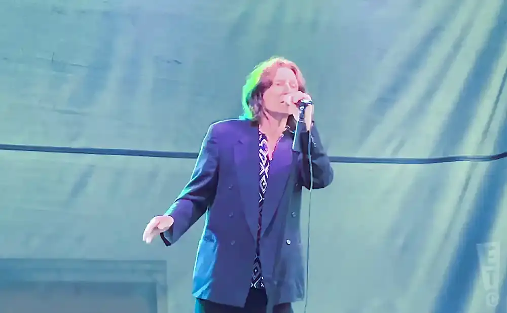 A man with brown hair and a patterned shirt sings into a microphone, wearing a dark suit jacket on stage.