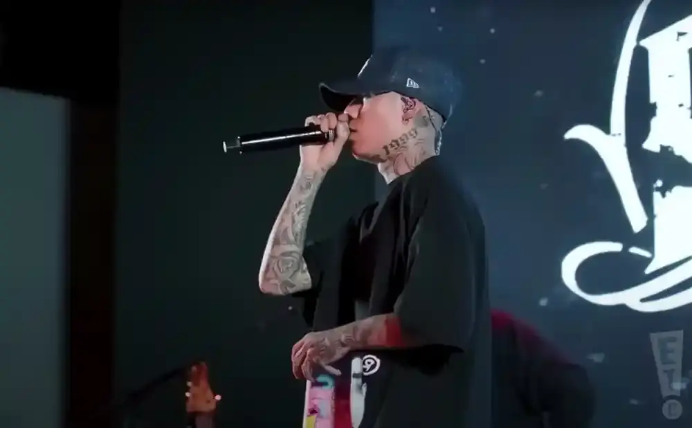 A man in a black t-shirt and baseball cap sings into a microphone on stage. He has tattoos on his neck and arms.