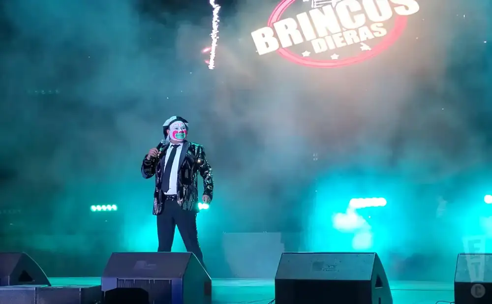A clown with face paint sings into a microphone on a stage lit with blue and green lights, smoke fills the air.