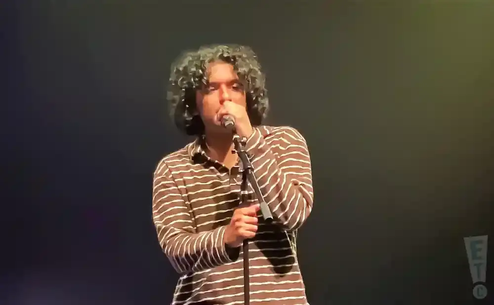 A person with curly hair sings into a microphone while wearing a brown and white striped shirt.