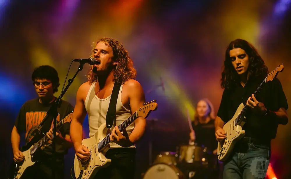 A band performs on stage with colorful lighting. The lead singer is in focus, singing into a microphone and playing a white guitar.