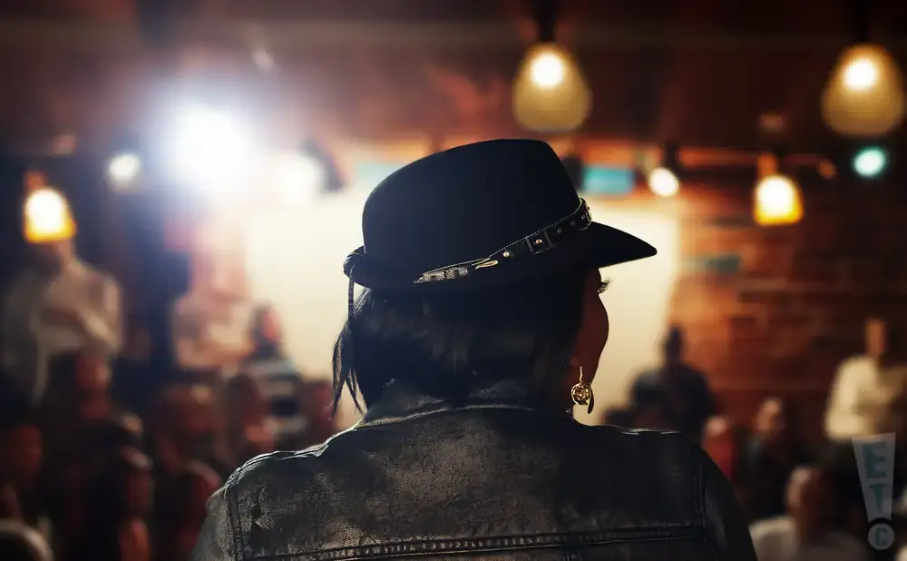 Woman in black hat and leather jacket seen from behind at a performance.